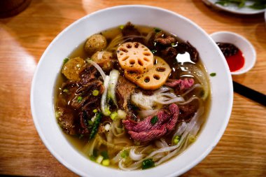 Bir kase çorba, erişte ve et. Kâse beyaz ve içinde kaşık var.