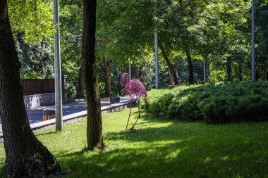 Pembe flamingo heykeli, telden örülmüş, yüksek bacaklar, çimenlerin üzerinde duruyor, park, ağaçların arasında, orijinal, şehir, yürüyüş, seyahat, manzara, doğa, gezinti, yaz, bahar, çocuk kampı, turistler, dinlenme, 