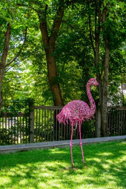 Pembe flamingo heykeli, telden örülmüş, yüksek bacaklar, çimenlerin üzerinde duruyor, park, ağaçların arasında, orijinal, şehir, yürüyüş, seyahat, manzara, doğa, gezinti, yaz, bahar, çocuk kampı, turistler, dinlenme, 
