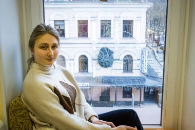 Genç kız pencerenin kenarında oturur ve sanat stüdyosuna, sokağa, eve, kışa gülümser.