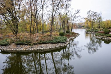 Sahildeki ağaçlar ve çalılar, taş kıyılar, köprü, nehir, göl, suya yansıyan ağaçlar, sarı-kırmızı, yapraklar, renkler, bulutlar, sonbahar, modern manzara,