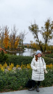 Beyaz yağmurluk ve şapkalı bir kadın köprünün kenarındaki ağaçlar sonbahar parkındaki suyun yanındaki portakal sarısı çalılar göller, yeşillik, manzara, dekoratif...,