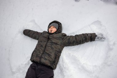 Sekiz yaşında bir çocuk karda yatıyor, bir kar meleği yapıyor, gülümsüyor, kırmızı yanaklar, karla kaplı eldivenler, şapka, buz, kış, eğlence, yürüyüş, dinlenme, hafta sonu