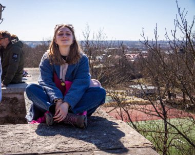 Genç kız, merceğe bakıyor, memnun, oturuyor, bacak bacak bacak üstüne atıyor, güneş gözlüğü takıyor, çitlerde, gözlem güvertesinde, şehrin üzerinde, sarışın, gülümseyen, panorama, üst manzara, parklar, ufuk, Avrupa, tepeler, turist, çıplak ağaçlar, kadın, doğa, gökyüzü, seyahat