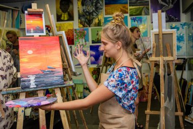 Genç bir kız, sarışın, önlüklü, tuvale, fırçaya, sehpaya, deniz manzarasına, bota, bir sanat atölyesine, el işine, usta sınıfına, duvarlara, arka planda dekora resim çiziyor.