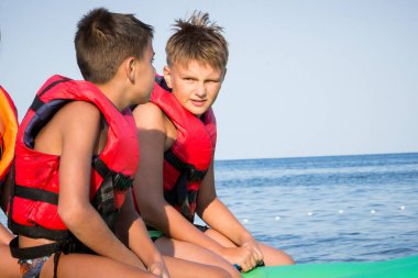 İki adam şişme bir yatakta, suyun üzerinde, bronzlaşmış bedenler, can yelekleri, sırtlar, güneş, ufuk, durgun yaz, seyahat, tatil, gençler., 