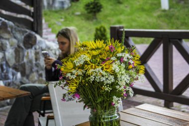 large bouquet wildflowers, standing  glass jar, on table, cafe, blond girl, sitting at table, foreground, looking at phone, multi-colored, yellow daisies