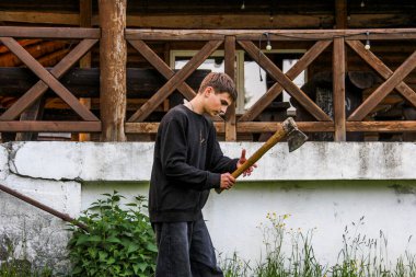 Genç bir çocuk demir bir baltayla dalları kesiyor, yan manzara, tam boy, ahşap bir evin yanında, siyah paita, kot pantolon, kamp ateşi için odun, çayır, akşam, yaz, orman, çocuk kampı.