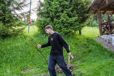 Elinde demir baltası olan genç bir çocuk, ateş için odun, ön manzara, boy, ahşap bir evin yanında, siyah pantolon, kot pantolon, çayır, akşam, yaz, orman, çocuk kampı.