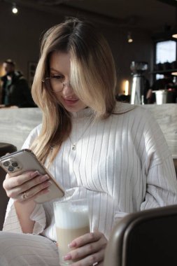 Kafede gözlüklü, latte içen ve baştan çıkarıcı bir şekilde gülümseyen genç ve güzel bir kadın.