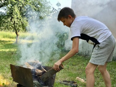 12 yaşındaki bir çocuk bahçedeki ızgaraya odun koyar.