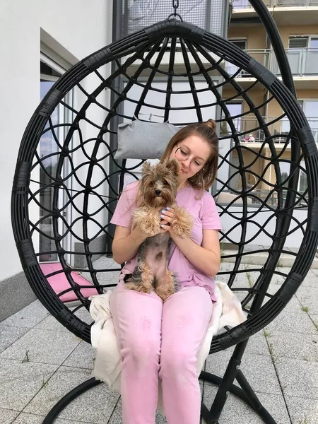 眼鏡をかけ ピンクの服を着ている若い女性が 彼女の腕に小さなヨークの犬を抱えて 彼と遊んでいるコクーンチェアのテラスに座っている — ストック写真