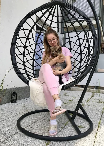 A young woman in glasses and wearing pink clothes is sitting on the terrace in a cocoon chair holding a small Yorkie dog in her arms and playing with him