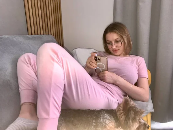 一个戴着眼镜 穿着粉色衣服的女孩躺在沙发上 手里拿着电话 — 图库照片