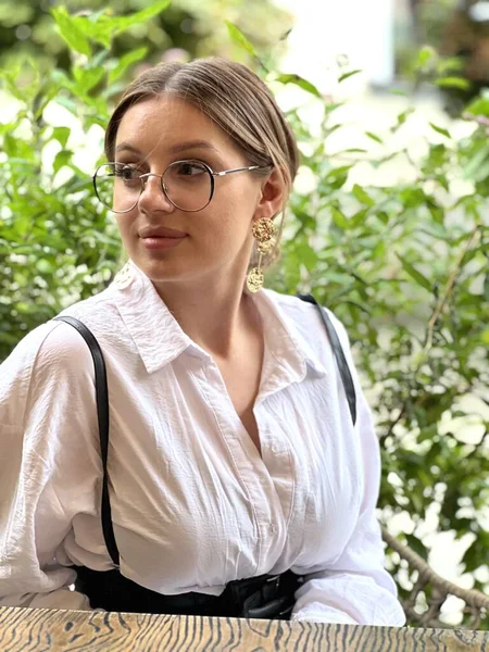 a young woman in glasses and a white blouse sits outside at a table in a cafe and waits for an appointment