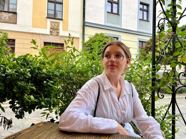a young woman in glasses and a white blouse sits outside at a table in a cafe and waits for an appointment