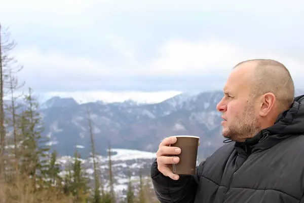 A middle-aged man drinks coffee against the background of snow-capped mountains