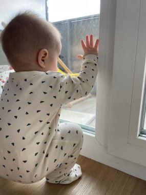 Balkon penceresinin yanında duran küçük bir çocuk yüksek binaların bahçesine bakıyor.