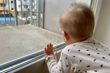 Balkon penceresinin yanında duran küçük bir çocuk yüksek binaların bahçesine bakıyor.