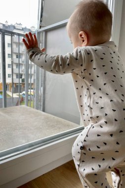 Balkon penceresinin yanında duran küçük bir çocuk yüksek binaların bahçesine bakıyor.