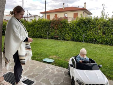 Küçük bir çocuk büyük beyaz bir bebek arabasıyla dolaşırken genç bir anne ona göz kulak oluyor.