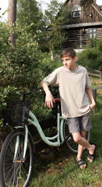 Bir genç, kırsal kesimde bir bisikletin yanında duruyor. Eski ahşap bir evin arka planına karşı.