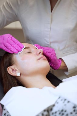 Pembe eldivenli bir güzellik uzmanı bir kızın yüzündeki nemlendirici maskeyi süngerle siler.