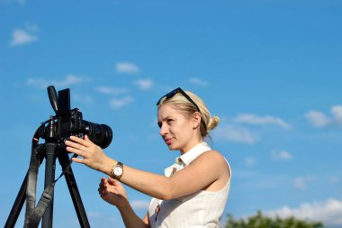 Mavi gökyüzünün arka planında güzel sarışın bir fotoğrafçı fotoğraf ve video çekmek için kamerayı ayarlıyor.