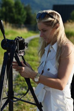Mavi gökyüzünün arka planında güzel sarışın bir fotoğrafçı fotoğraf ve video çekmek için kamerayı ayarlıyor..