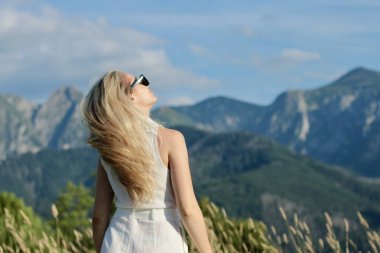 Beyaz takım elbiseli güzel sarışın bir kadın tarladaki dağların arkasında poz veriyor ve dinleniyor.