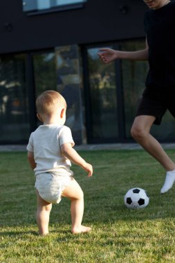 Bir ağabey ve bez takmış küçük bir çocuk bahçedeki yeşil çimlerin üzerinde top oynuyorlar.