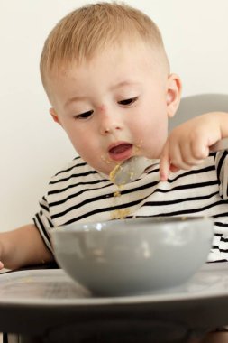 Küçük ciddi bir çocuk yüksek bir sandalyede otururken kaşıkla bağımsız olarak yemek yer..