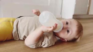 Yerde yatan küçük bir çocuk elinde bir bebek şişesi tutuyor ve ondan süt içiyor.
