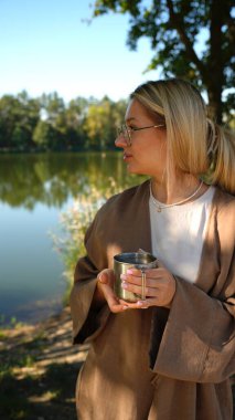 Nehir ve ormanın mavi arka planında kahve ya da çay içen güzel genç bir kadın. Manzarası güzel.