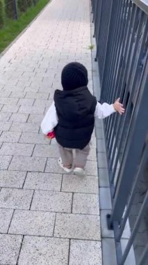 Küçük bir çocuk yolda yürüyor, demir bir çitin yanında tek başına.