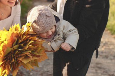 Kollarında küçük bir çocuk olan genç bir aile, elinde bir buket sarı akçaağaç yaprağıyla parktan mutlu bir şekilde uzaklaşıyor ve çocuğa gülümsüyor.