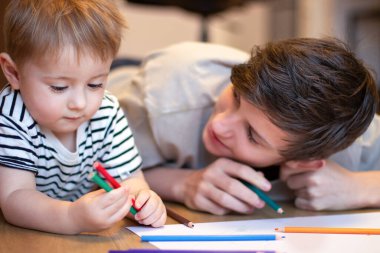 İki çocuk renkli kalemlerle resim çiziyor. Bir genç ve bir küçük kardeş yerde yatarken silah çekerler..