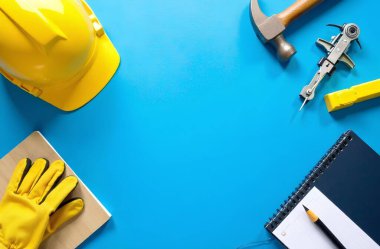 Eldivenli sarı kask, defter, kalem ve aletler mavi arka planda..