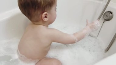 Çocuk suda köpükle banyo yapar, bir oyuncakla oynar, musluktan akan eliyle su tutar.