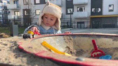 İki yaşında bir çocuk, oyun parkında oyuncak arabasıyla oyun oynuyor, çocuk ilkbaharın başında sıcak kıyafetler giyiyor.