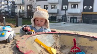 İki yaşında bir çocuk, oyun parkında oyuncak arabasıyla oyun oynuyor, çocuk ilkbaharın başında sıcak kıyafetler giyiyor.