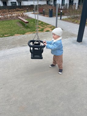 Küçük bir çocuk salıncakta mutlu bir şekilde sallanıyor, çocuk sıcak mavi bir ceket ve bej bir şapka giyiyor.