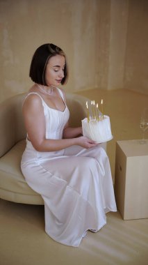 Kısa saçlı, beyaz elbiseli, elinde doğum günü pastası ve doğum günü için mumlar tutan tatlı bir genç kadın.