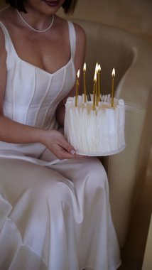 Kısa saçlı, beyaz elbiseli, elinde doğum günü pastası ve doğum günü için mumlar tutan tatlı bir genç kadın.
