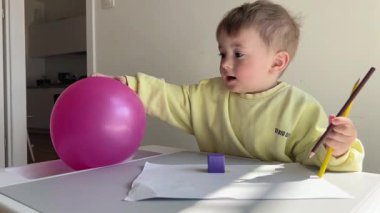 Küçük bir çocuk masada oturuyor ve kalemler ve pembe bir balonla oynuyor.