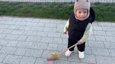 Bir çocuk bahçede bir patikada yürüyor, tahta bir oyuncakla oynuyor.