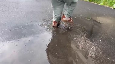 Kahverengi ayakkabılı küçük bir çocuk bahar yağmurundan sonra ayaklarını yere vurarak su sıçratıyor.