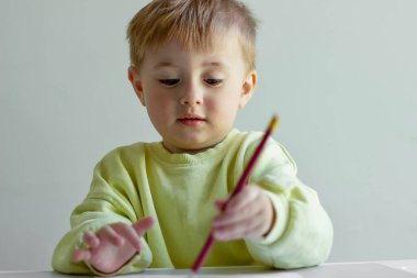 Sarı kazaklı neşeli küçük çocuk masada renkli kalemlerle kağıt üzerine resim çiziyor.