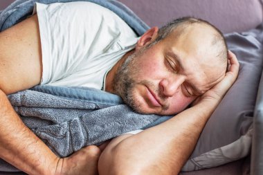 Yorgun ve yorgun orta yaşlı bir adam gri bir battaniyenin altında uyuyor.