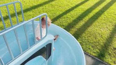 Küçük bir çocuk topun peşinden mavi bir sarmal kaydırağına gidiyor. Bir çocuk çam ormanının yakınındaki bir parkta oyun oynuyor.